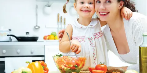 Mama i córka przygotowują sałatkę. W kuchni widać warzywa, oliwę i chleb. To zdrowa żywność, bogata w oligosacharydy, które wspierają florę bakteryjną.