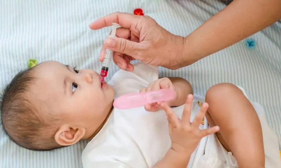 Dłoń podaje niemowlęciu witaminę D z pipetki. Dziecko leży na boku, ubrana w białą koszulkę.