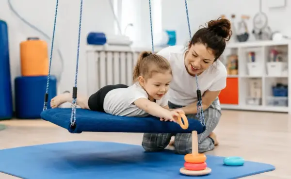 Dziecko na huśtawce podczas terapii integracji sensorycznej co to jest. Terapeuta pomaga dziecku w zabawie.